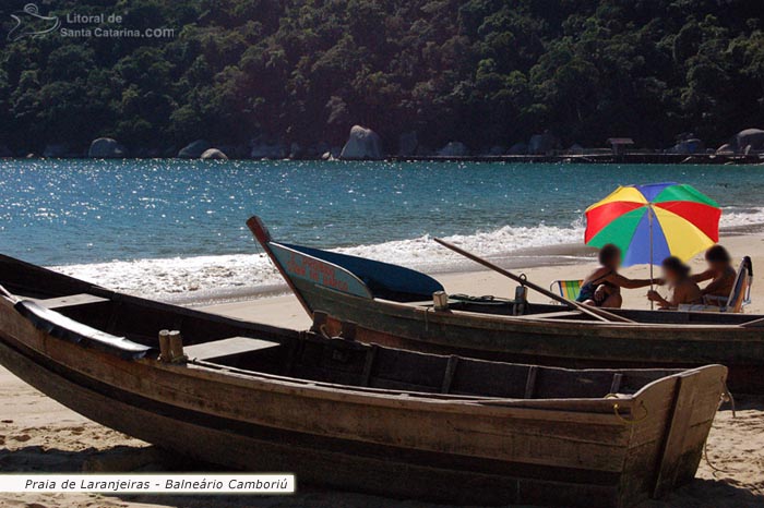 Barco parado nas areias da praia de laranjeiras em balneário camboriú e ao fundo famílias batendo um papo em baixo do guarda sol.