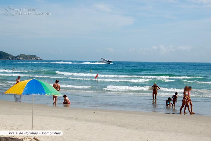 Crianças se divertindo na areia da praia de bombas e ao fundo uma lancha passando, formando assim um cenário maravilhoso.