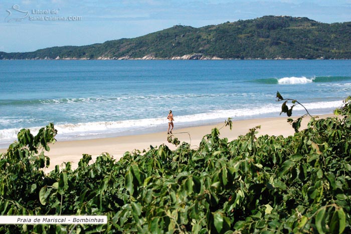 Moça correndo na Praia de Mariscal em Bombinhas e ao fundo um cenário maravilhoso de preservação a mata nativa.